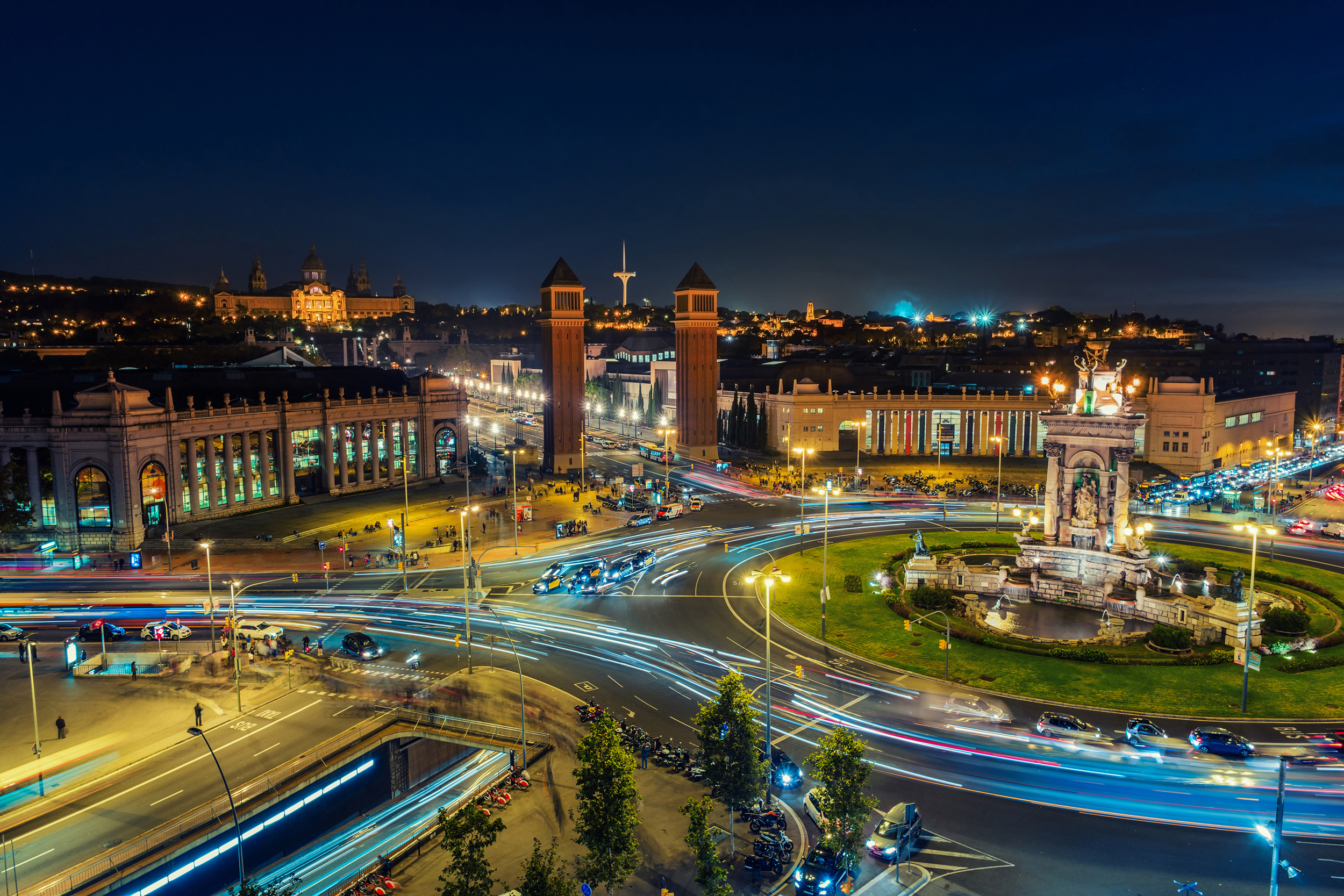 Spanish Square aerial view in Barcelona, Spain at night. This is the famous place with traffic light trails, fountain and Venetian towers, and National museum at the background. Blue sky