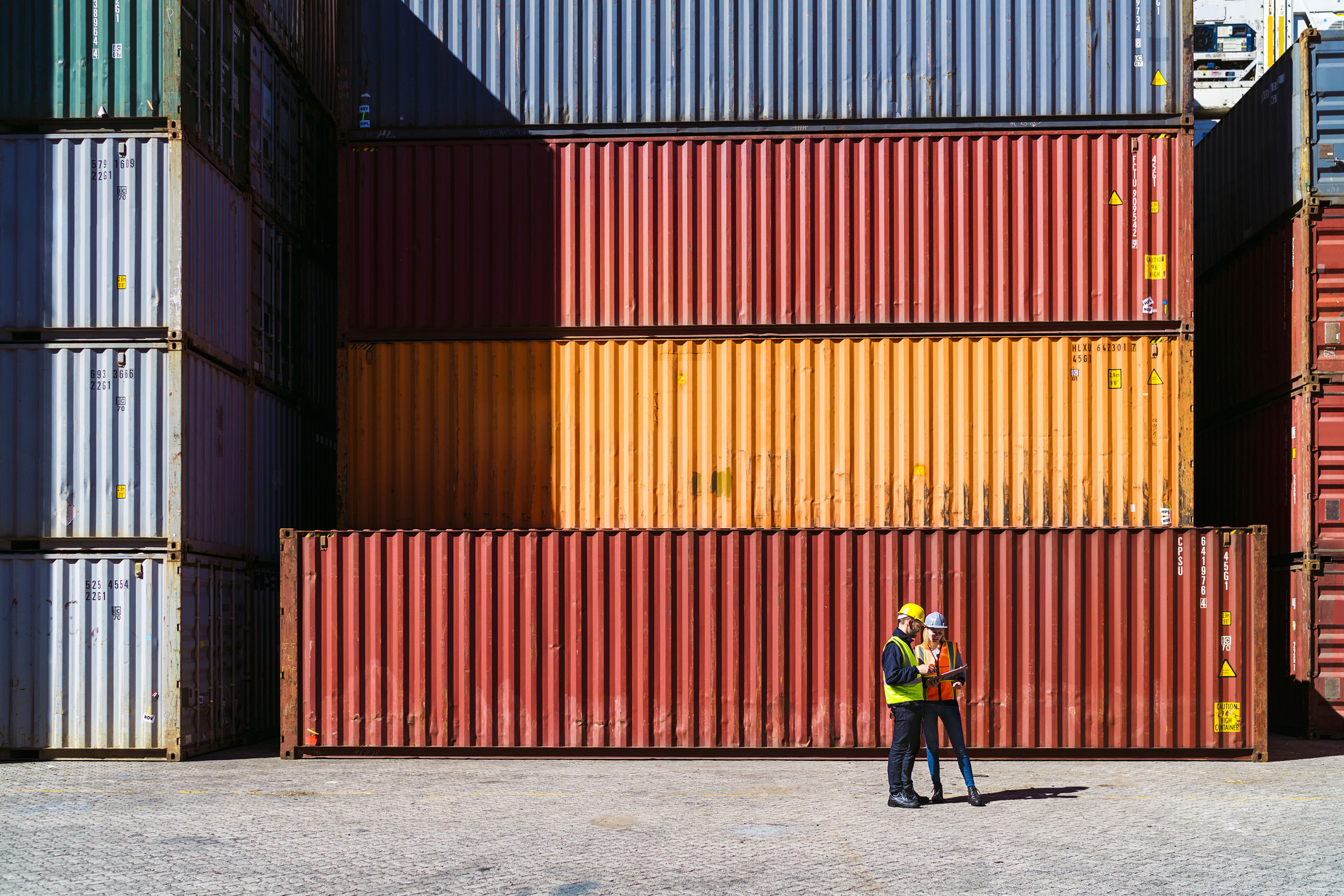 A photo of male and female dock workers having discussion. Full length of manual workers standing against cargo containers. They are working in shipping yard.