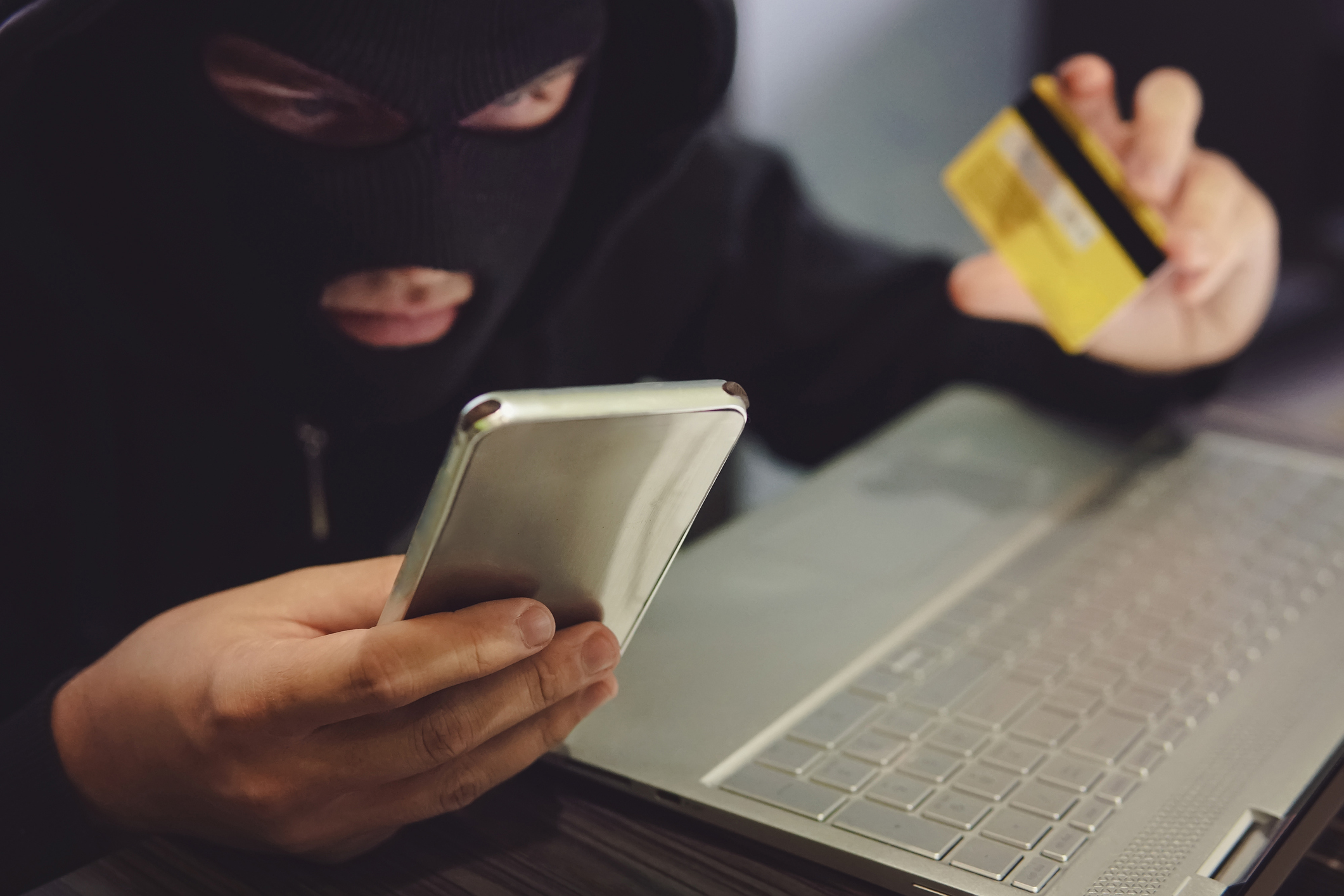 Male hacker in a robber mask uses phone, credit card and laptop in some fraudulent scheme. Cyber thief stole the personal data and credit card information. Hacker uses malware to steal user's money