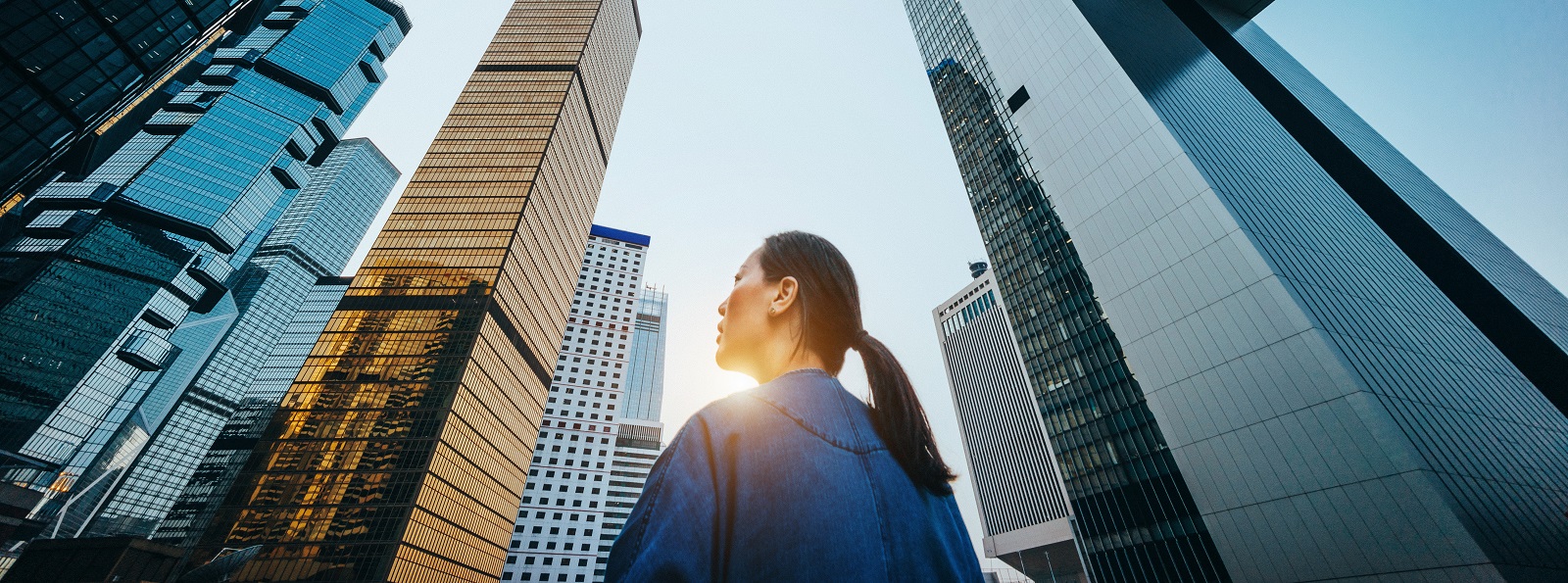 Low angle view of woman standing against modern skyscrapers and blue sky in city at sunrise