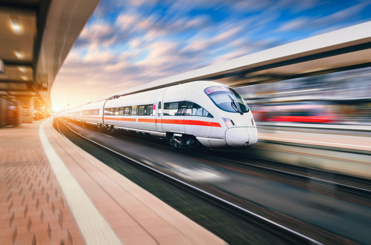 White modern speed train in motion on railway station at sunset. Train on railroad track with motion blur effect in Europe in evening. Railway platform. Industrial landscape. Railway tourism