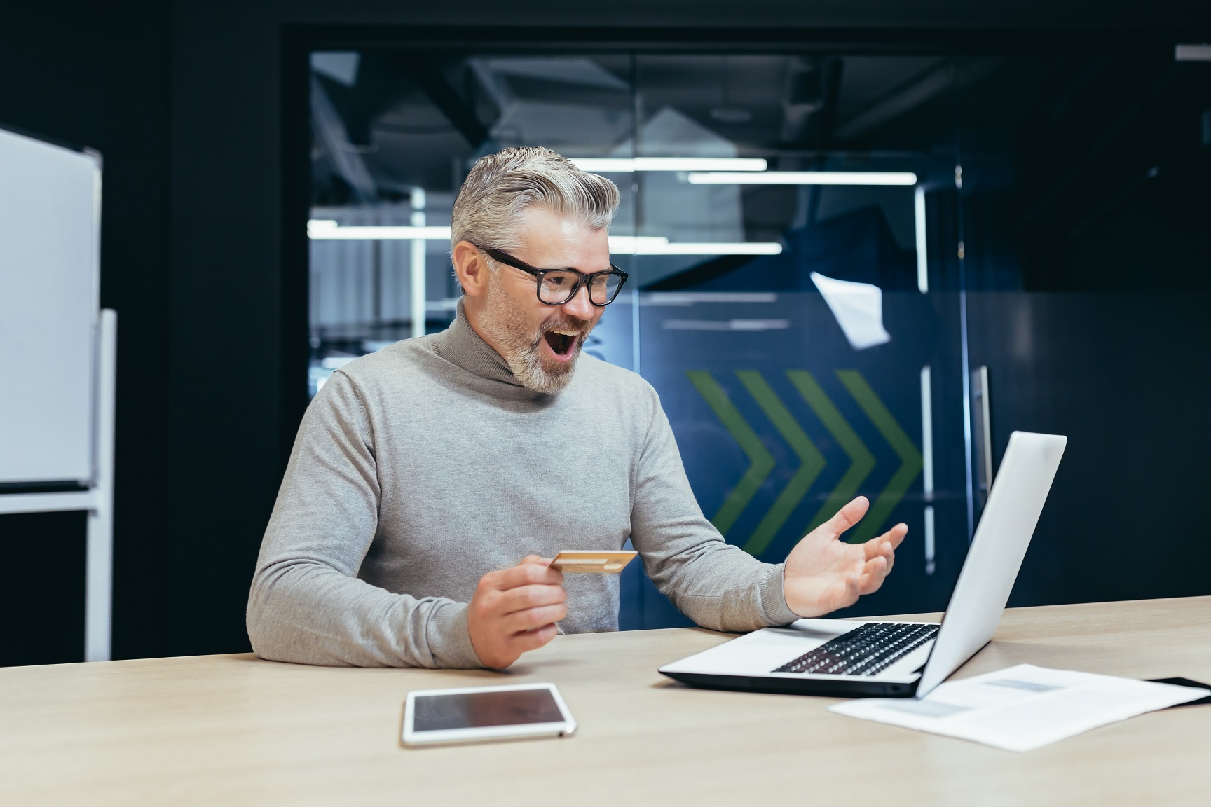 Happy and smiling businessman in office making bank transfer and online purchase in online store, mature man holding bank credit card and using laptop, happy with choice