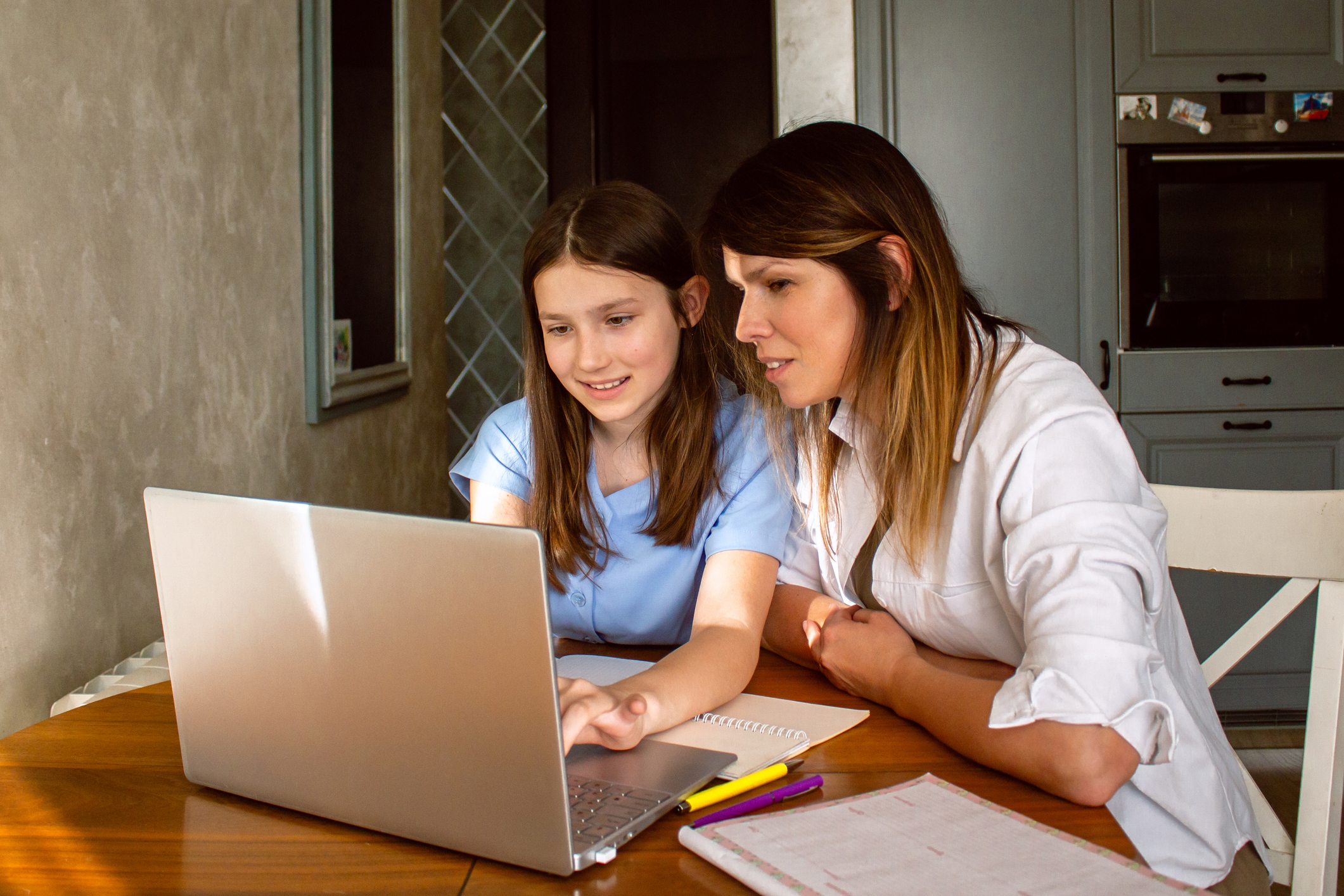 Daughter Showing Mom Something Exciting On Internet On Laptop Screen Sitting At Table In Kitchen
