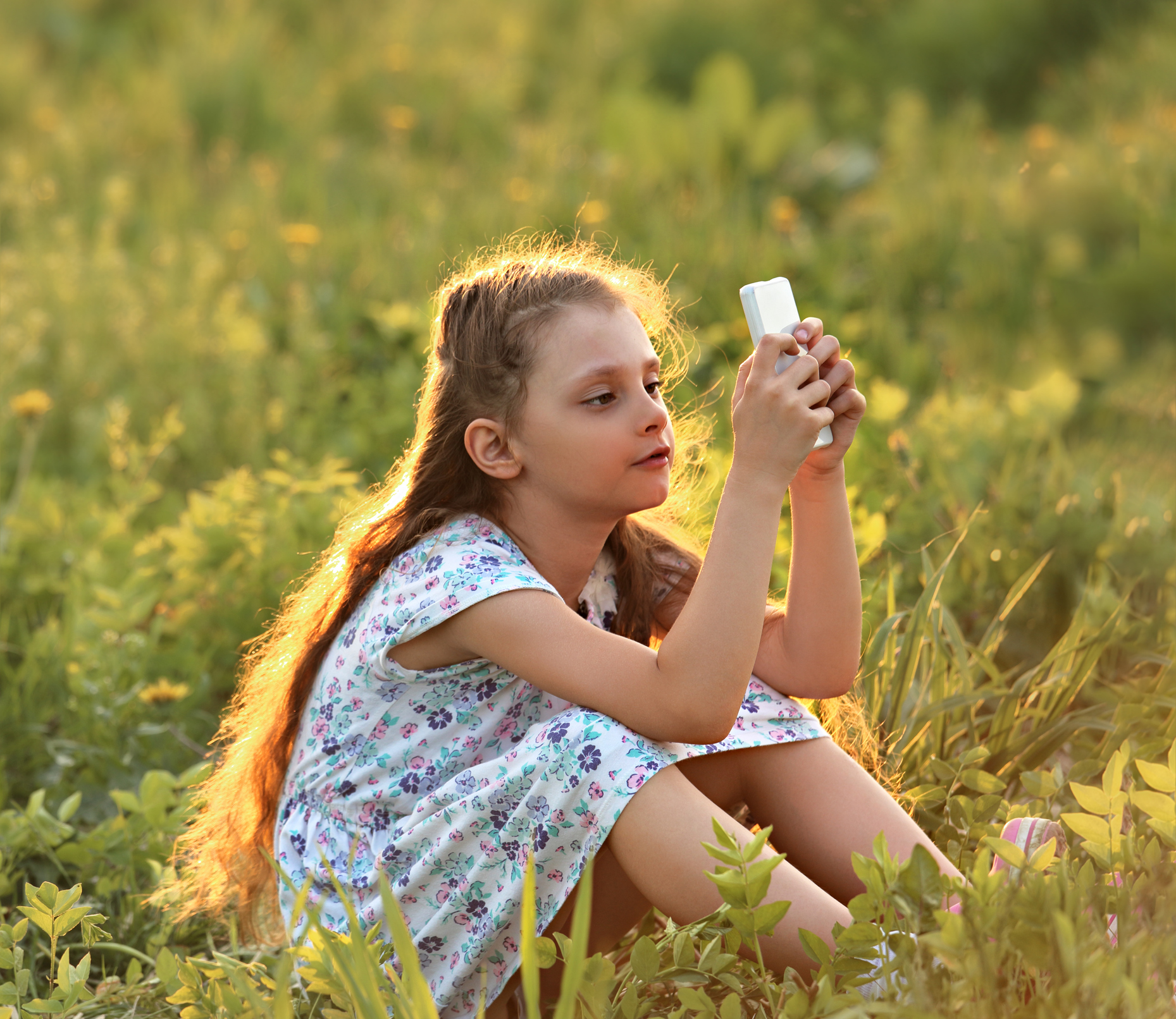Cute kid girl sitting on the glass and playing on mobile phone with serious face on summer background
